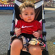Jariyah is registered to the contest to win money with this photo: toddler, child, stroller, curly_hair, red_shirt, nike_logo, plaid_shorts, outdoor, carnival, amusement_park, food_stand, lights, serious_expression, sunlight, pavement, seatbelt, summer, casual_clothing, young_child, portrait