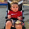 toddler, child, stroller, curly_hair, red_shirt, nike_logo, plaid_shorts, outdoor, carnival, amusement_park, food_stand, lights, serious_expression, sunlight, pavement, seatbelt, summer, casual_clothing, young_child, portrait