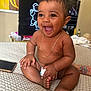 baby, smiling, laughing, sitting, diaper, gold_chain, indoor, bedspread, smartphone, tattoo, arm, person, child, cute, happy, skin, hair, face, expression, furniture