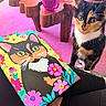 cat, calico_cat, painting, colorful_art, pink_carpet, wooden_table, pink_cup, flower_pattern, animal_portrait, indoor, pet, curious, artwork, sitting, closeup, decor, vibrant_colors, feline, domestic_cat, cozy