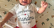 Malakai is registered to the contest to win money with this photo: baby, child, smiling, happy, infant, clothing, shirt, shorts, red_shorts, white_shirt, text_on_clothing, animal_print, bedding, patterned_sheet, lying_down, hand_raised, face, cute, indoors, person