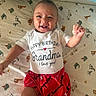 baby, child, smiling, happy, infant, clothing, shirt, shorts, red_shorts, white_shirt, text_on_clothing, animal_print, bedding, patterned_sheet, lying_down, hand_raised, face, cute, indoors, person