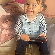 Joy a rejoint le concours — aidez-le/la à gagner de superbes lots ! toddler, child, smiling, remote_control, striped_shirt, overalls, boots, hair_clip, pillow, portrait_pillow, bed, blanket, indoor, cozy, furniture, happy, person, face, home, cute