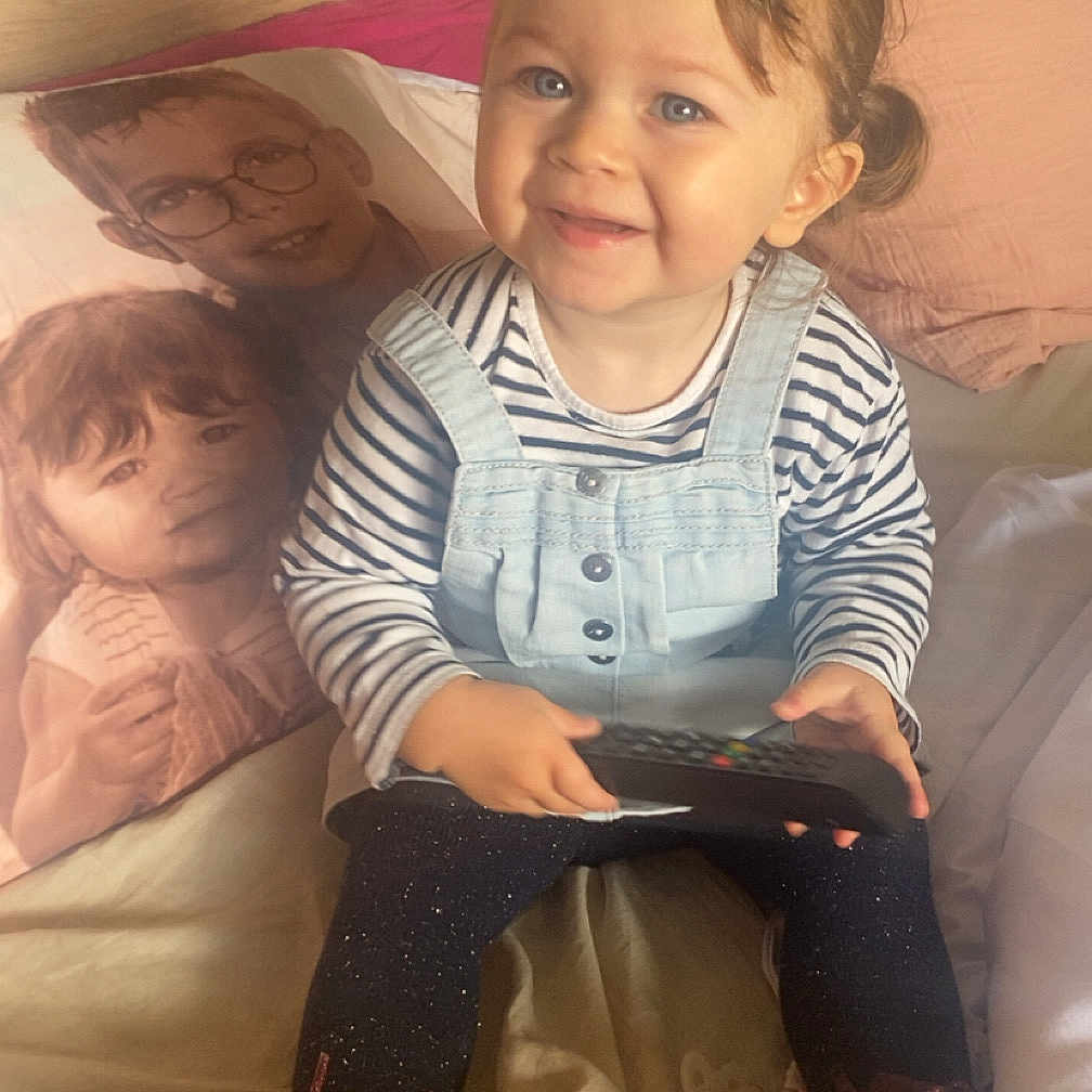 Joy a rejoint le concours — aidez-le/la à gagner de superbes lots ! bed, blanket, boots, child, cozy, cute, face, furniture, hair_clip, happy, home, indoor, overalls, person, pillow, portrait_pillow, remote_control, smiling, striped_shirt, toddler