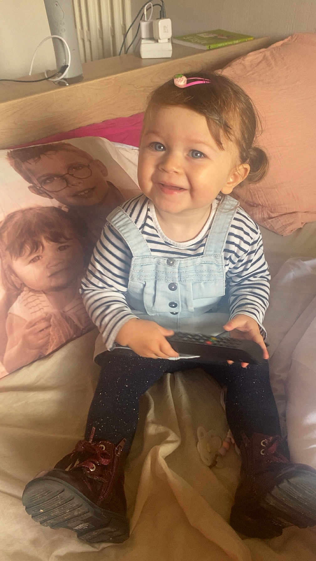 Joy a rejoint le concours — aidez-le/la à gagner de superbes lots ! toddler, child, smiling, remote_control, striped_shirt, overalls, boots, hair_clip, pillow, portrait_pillow, bed, blanket, indoor, cozy, furniture, happy, person, face, home, cute