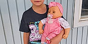 Remi is registered to the contest to win money with this photo: casual_clothing, child, concrete, daylight, doll, expression, face, footwear, hair, holding, outdoor, pacifier, person, shorts, standing, toy, tshirt, wall, window, young