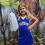 child, girl, cheerleader, pom_pom, blue_uniform, smile, long_hair, standing, hand_on_hip, indoor, horse_backdrop, cheerleading, portrait, yellow_trim, sportswear, confident, happy, background, floor, team_spirit