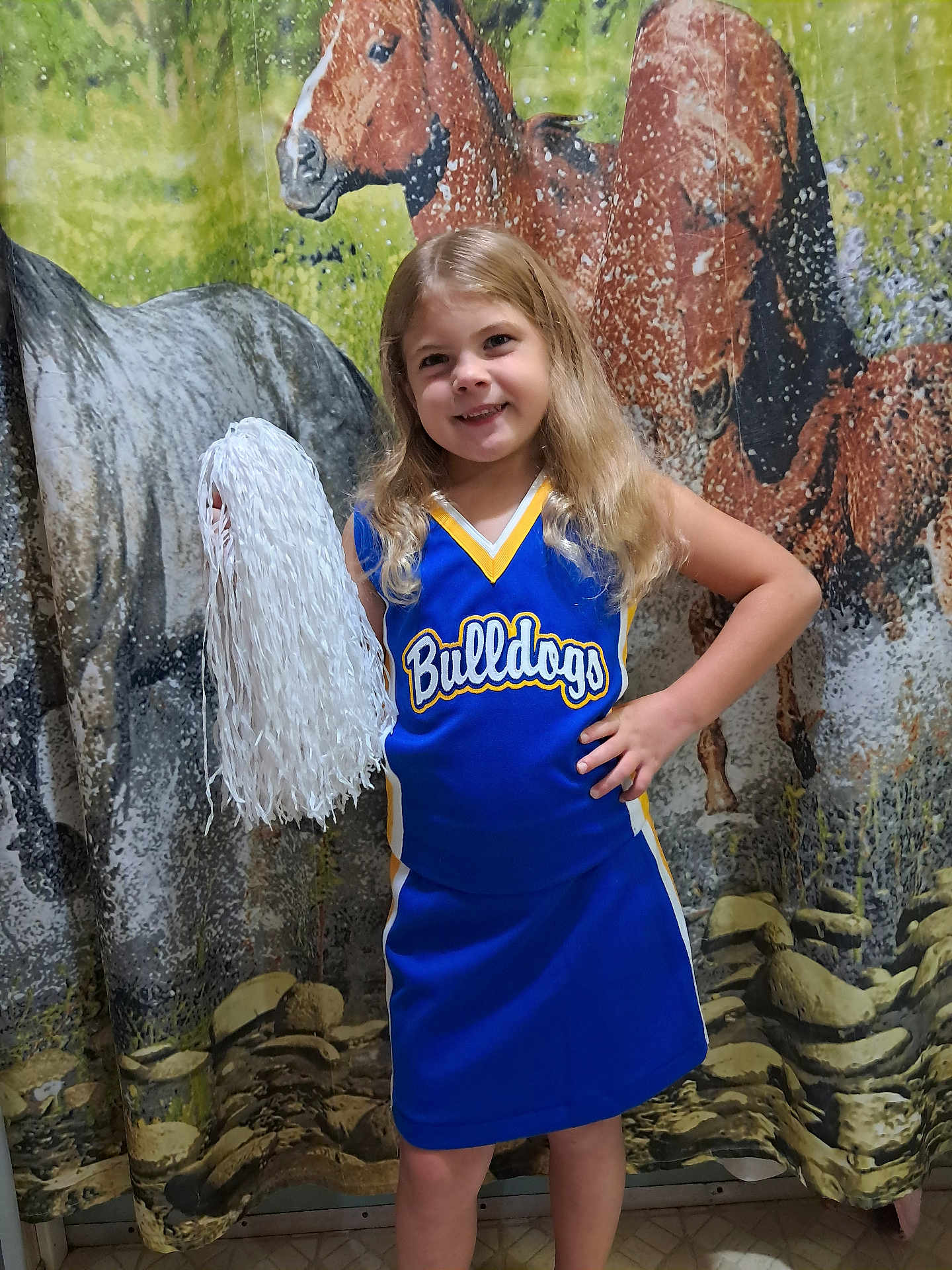 Remi is registered to the contest to win money with this photo: child, girl, cheerleader, pom_pom, blue_uniform, smile, long_hair, standing, hand_on_hip, indoor, horse_backdrop, cheerleading, portrait, yellow_trim, sportswear, confident, happy, background, floor, team_spirit