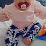 baby, child, pacifier, headband, bow, pink_sweater, black_and_white_pants, sitting, bed, toy, indoor, cute, infant, foot, face, person, clothing, looking, curious, childrenswear