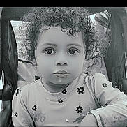 Ashanti is registered to the contest to win money with this photo: child, toddler, curly_hair, face, stroller, shirt, flower_pattern, food, expression, portrait, black_and_white, sitting, young_child, cute, eyes, mouth, clothing, indoor, baby, person
