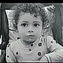 child, toddler, curly_hair, face, stroller, shirt, flower_pattern, food, expression, portrait, black_and_white, sitting, young_child, cute, eyes, mouth, clothing, indoor, baby, person