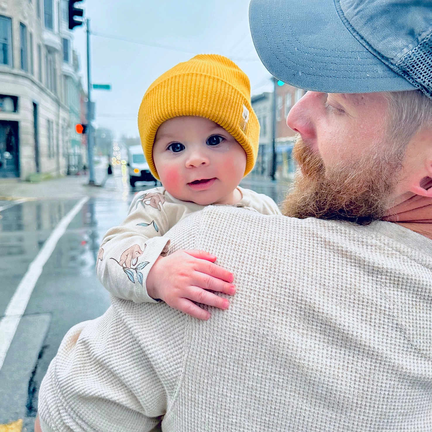 Oliver is registered to the contest to win money with this photo: baby, beanie, buildings, child, clothing, facial_hair, hand, holding, love, man, outdoor, person, portrait, rainy_weather, sidewalk, street, traffic_light, urban, warm_clothing, yellow_hat