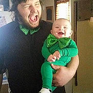 Micah is registered to the contest to win money with this photo: adult, baby, child, green_clothing, bow_tie, smiling, laughing, indoor, home, person, holding, joyful, casual, window, light, happy, family, portrait, celebration, fun