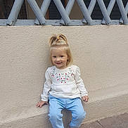Rebeca a rejoint le concours — aidez-le/la à gagner de superbes lots ! blonde_hair, blue_pants, casual_clothing, child, concrete_floor, cute, daylight, fashion, floral_embroidery, happy, outdoor, person, pink_shoes, pony_tail, sitting, smile, toddler, wall, white_top, young_child