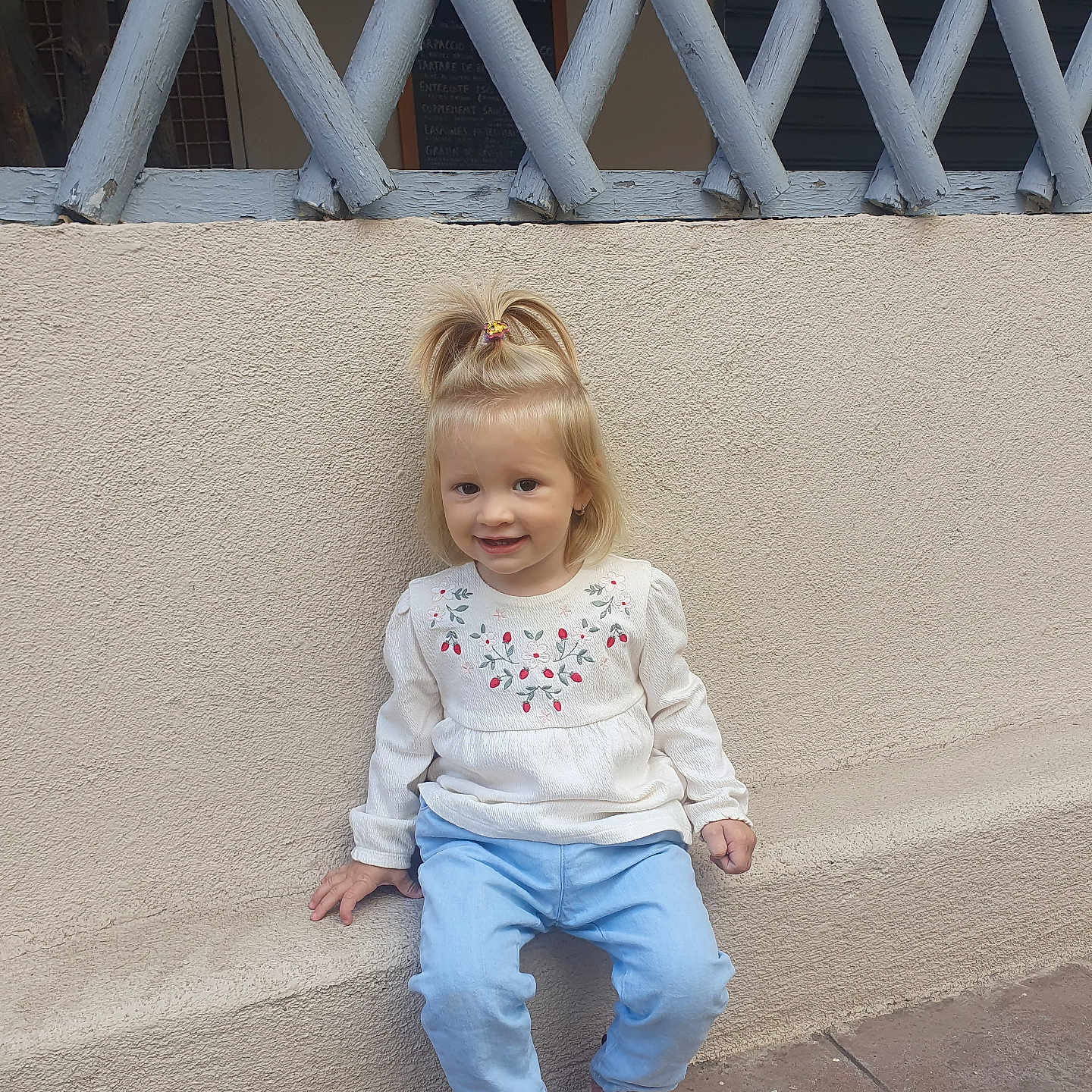 Rebeca a rejoint le concours — aidez-le/la à gagner de superbes lots ! blonde_hair, blue_pants, casual_clothing, child, concrete_floor, cute, daylight, fashion, floral_embroidery, happy, outdoor, person, pink_shoes, pony_tail, sitting, smile, toddler, wall, white_top, young_child