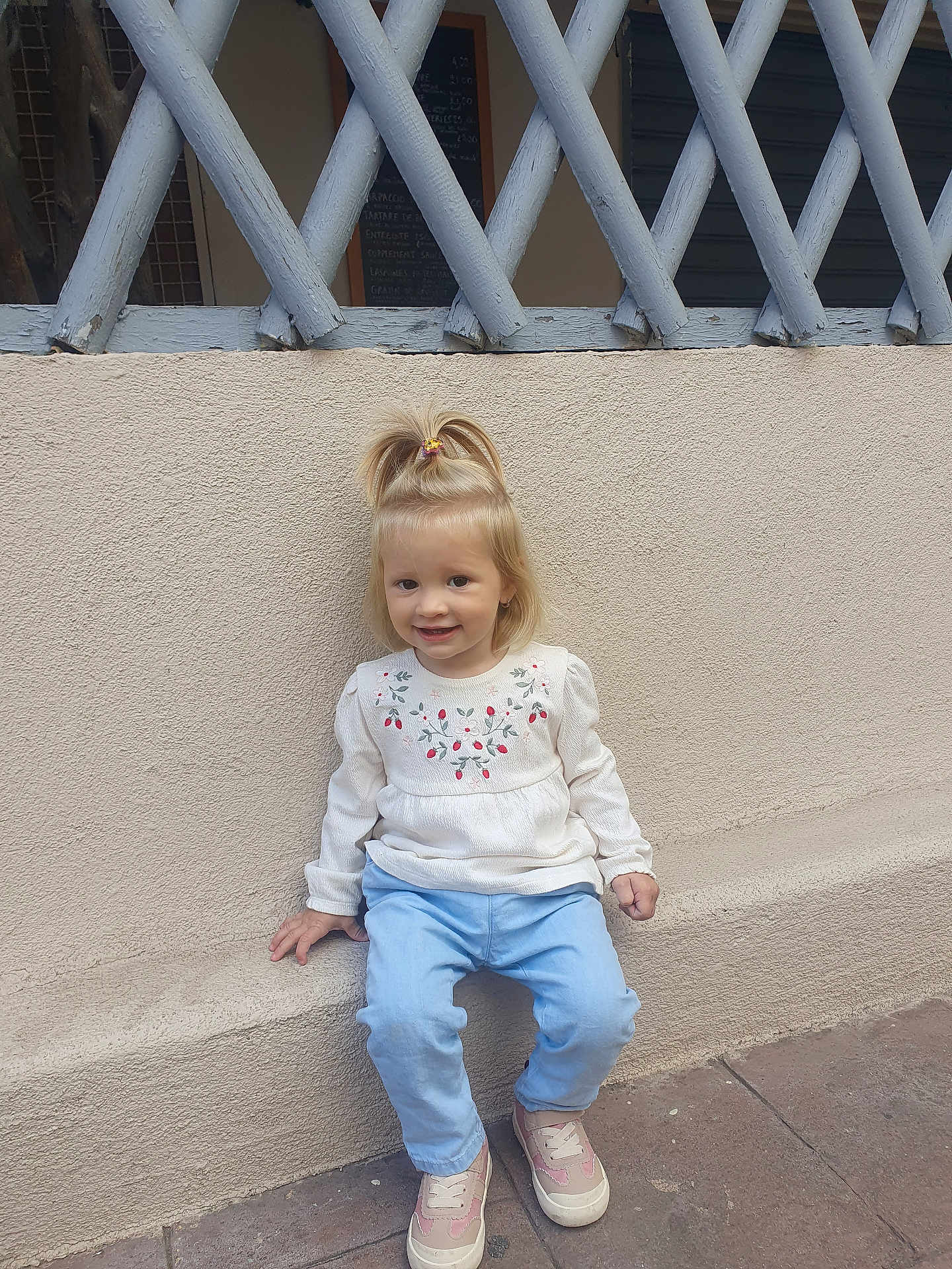 Rebeca a rejoint le concours — aidez-le/la à gagner de superbes lots ! blonde_hair, blue_pants, casual_clothing, child, concrete_floor, cute, daylight, fashion, floral_embroidery, happy, outdoor, person, pink_shoes, pony_tail, sitting, smile, toddler, wall, white_top, young_child