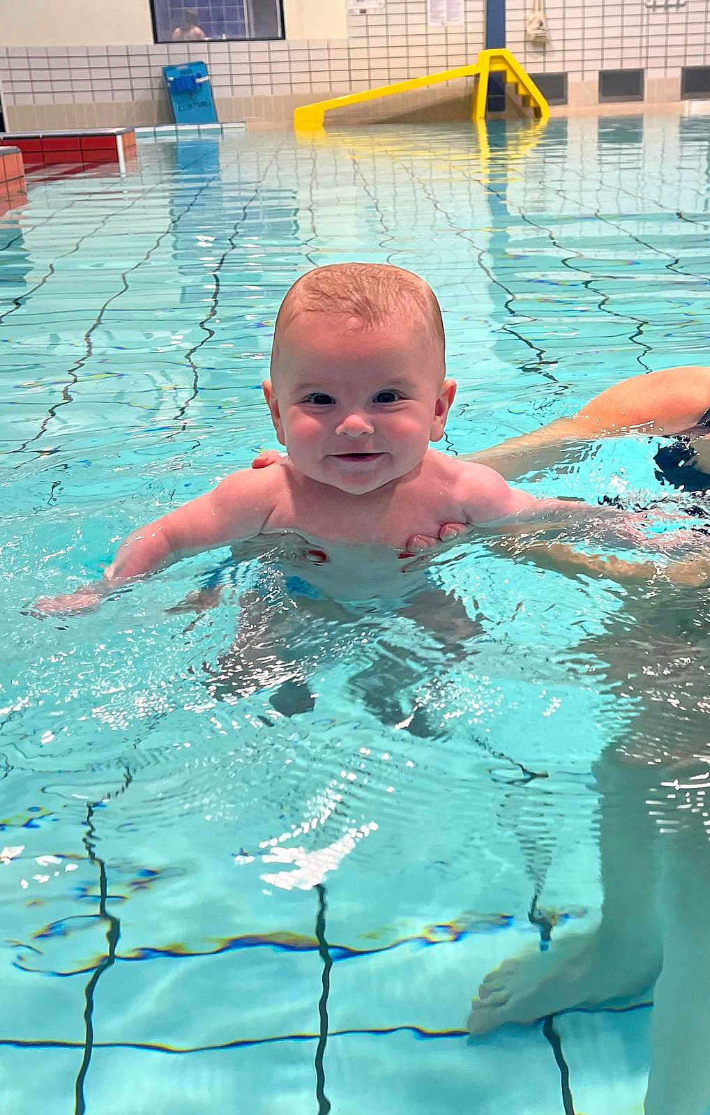 Clément participe au concours pour gagner de l'argent avec cette photo : baby, infant, swimming_pool, water, smile, happy, splash, indoor_pool, tiles, yellow_slide, parent_hand, supporting_hand, portrait, cute, floating, bare_chest, legs, reflection, waves, turquoise