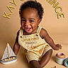 baby, child, toddler, smiling, happy, portrait, sitting, overalls, striped_clothing, curly_hair, dark_skin, toy_sailboat, sailboat, starfish, cups, beach_theme, studio, warm_lighting, text_overlay, fashion_photo