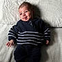baby, infant, smiling, tongue_out, sweater, striped_clothing, pants, lying_down, blanket, textured_blanket, cozy, cute, playful, child, person, indoor, happy, face, head, skin