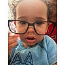 child, toddler, glasses, blue_shirt, cute, face, portrait, indoors, person, baby, expression, curly_hair, hand, closeup, clothing, eyewear, accessory, background, toy, hello_kitty