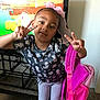 child, girl, peace_sign, pink_backpack, headband, cartoon_shirt, indoor, carpet, television, game_screen, standing, posing, smiling, cute, playful, person, clothing, accessory, home, fun
