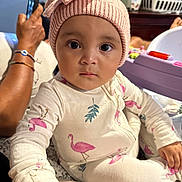 Hazel joined the competition — help win amazing prizes! baby, infant, child, portrait, sitting, indoors, pink_hat, bunny_hat, onesie, flamingo_pattern, wide_eyes, neutral_expression, toy, playpen, laundry_basket, adult_hand, smartphone, home, closeup, family