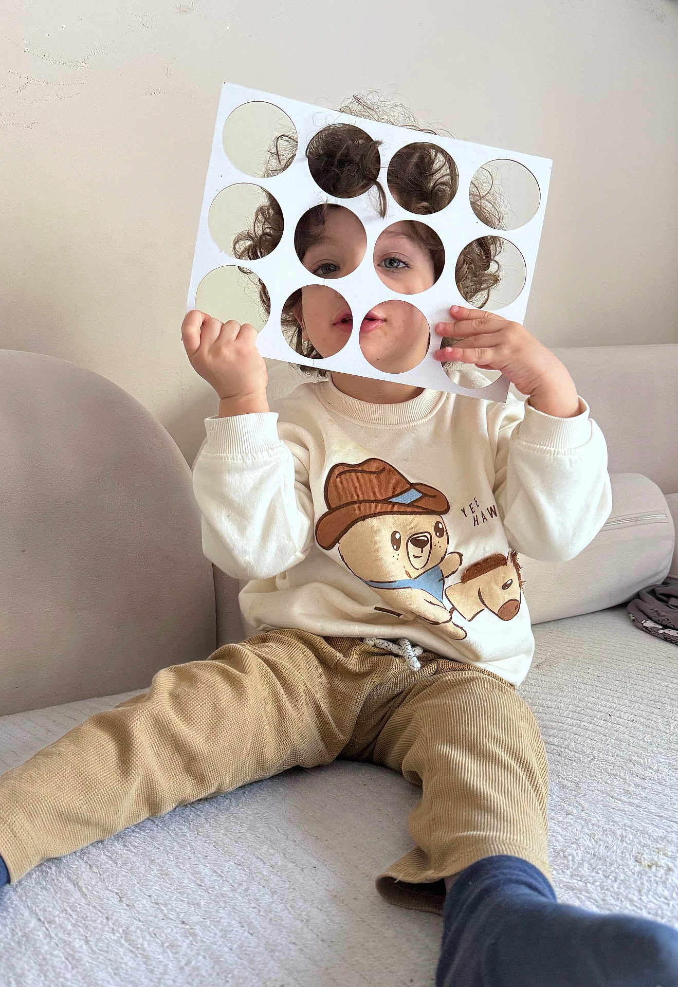 Zayn participe au concours pour gagner de l'argent avec cette photo : child, curly_hair, stencil, circular_cutouts, sweater, bear_graphic, yee_haw, pants, sitting, couch, indoor, playful, face, hands, light_background, neutral_colors, cozy, comfort, portrait, casual_clothing