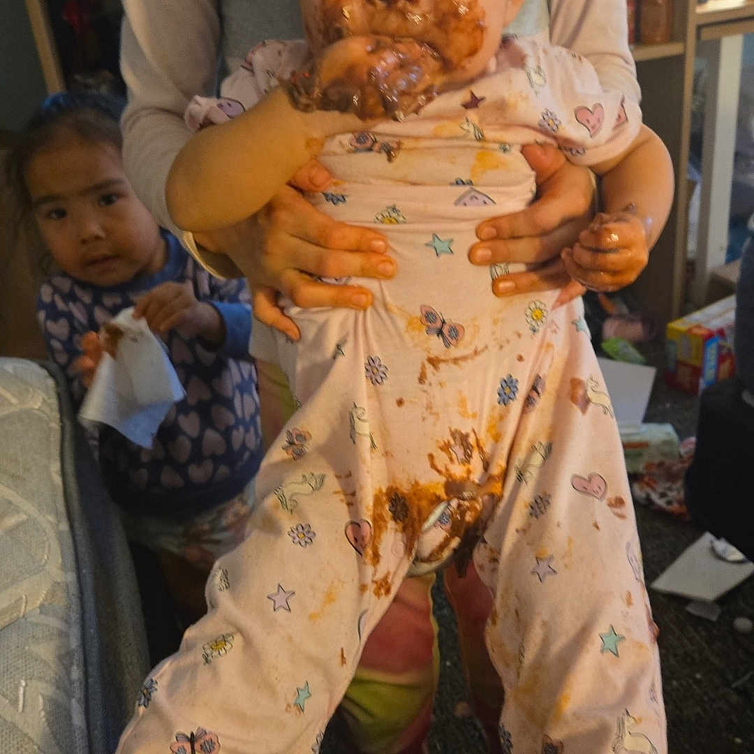 Shameka is registered to the contest to win money with this photo: adult_hands, baby, background, carpet, child, chocolate, clothing, face, floor, furniture, holding, indoor, living_room, messy, pajamas, person, sibling, smiling, tissue, toy