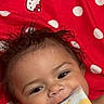 Naomi is registered to the contest to win money with this photo: baby, child, face, smiling, red_blanket, polka_dots, hello_kitty, lotion_packet, hands, happy, cute, tousled_hair, infant, lying_down, close_up, skin, chewing, background, soft_toys, home