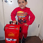 Salguero is registered to the contest to win money with this photo: child, boy, toddler, smiling, portrait, indoor, hallway, door, door_handle, red_clothing, hoodie, sweatpants, sneakers, luggage, suitcase, toy_car, race_car, cartoon, standing, happy