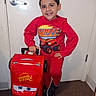 child, boy, toddler, smiling, portrait, indoor, hallway, door, door_handle, red_clothing, hoodie, sweatpants, sneakers, luggage, suitcase, toy_car, race_car, cartoon, standing, happy