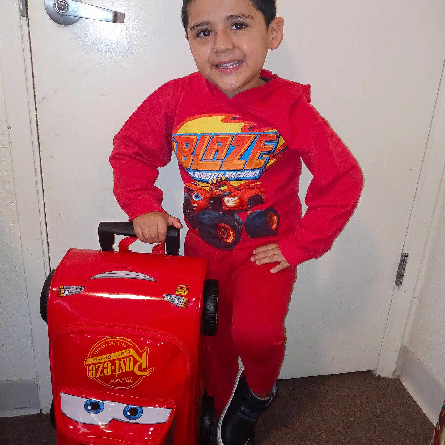 Salguero is registered to the contest to win money with this photo: child, boy, toddler, smiling, portrait, indoor, hallway, door, door_handle, red_clothing, hoodie, sweatpants, sneakers, luggage, suitcase, toy_car, race_car, cartoon, standing, happy