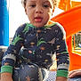 child, toddler, playground, slide, orange, blue, pajamas, dinosaur_pattern, curly_hair, hand, face, outdoor, sunlight, daylight, play, clothing, person, young_child, expression, park