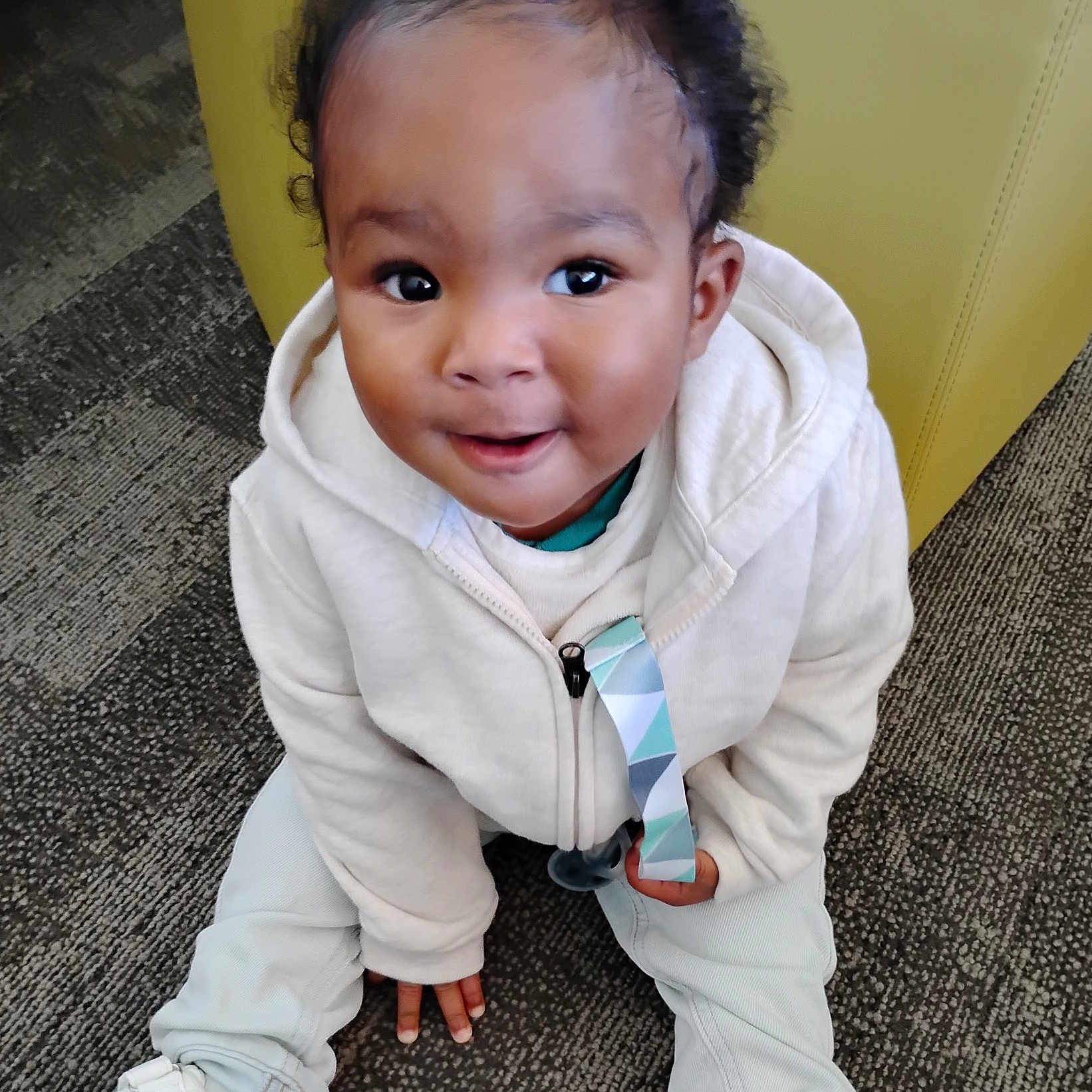 Veyquan joined the competition — help win amazing prizes! baby, child, infant, smiling, portrait, sitting, indoors, white_clothing, hoodie, jacket, pants, shoes, curly_hair, face, eyes, carpet, chair, yellow_background, colorful_strap, happy