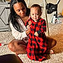 baby, woman, indoor, carpet, red_clothing, plaid_pattern, smiling, sitting, supporting, furniture, television, cabinet, floor, person, home, light, face, child, holding, curious
