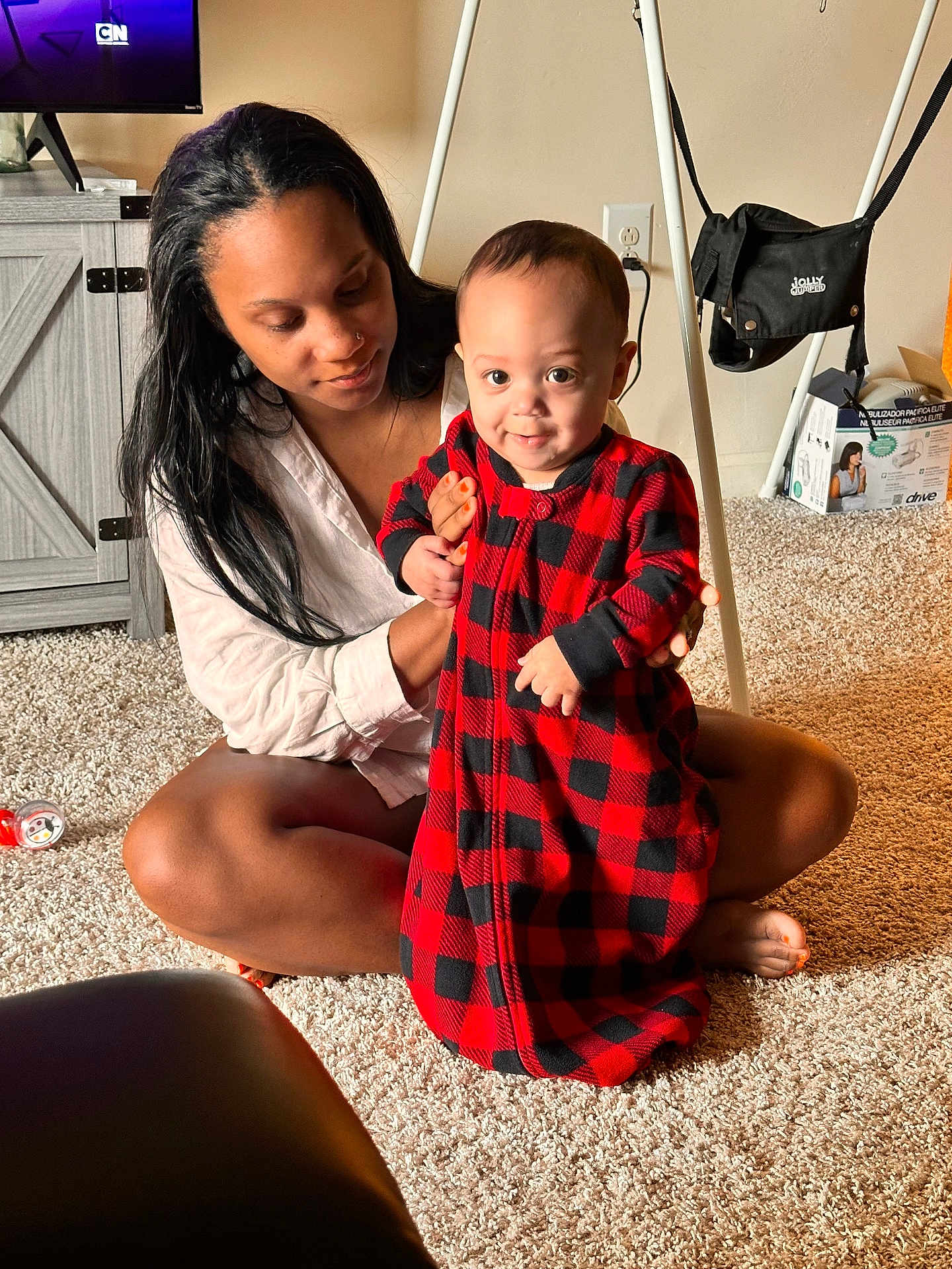 Christopher joined the competition — help win amazing prizes! baby, woman, indoor, carpet, red_clothing, plaid_pattern, smiling, sitting, supporting, furniture, television, cabinet, floor, person, home, light, face, child, holding, curious