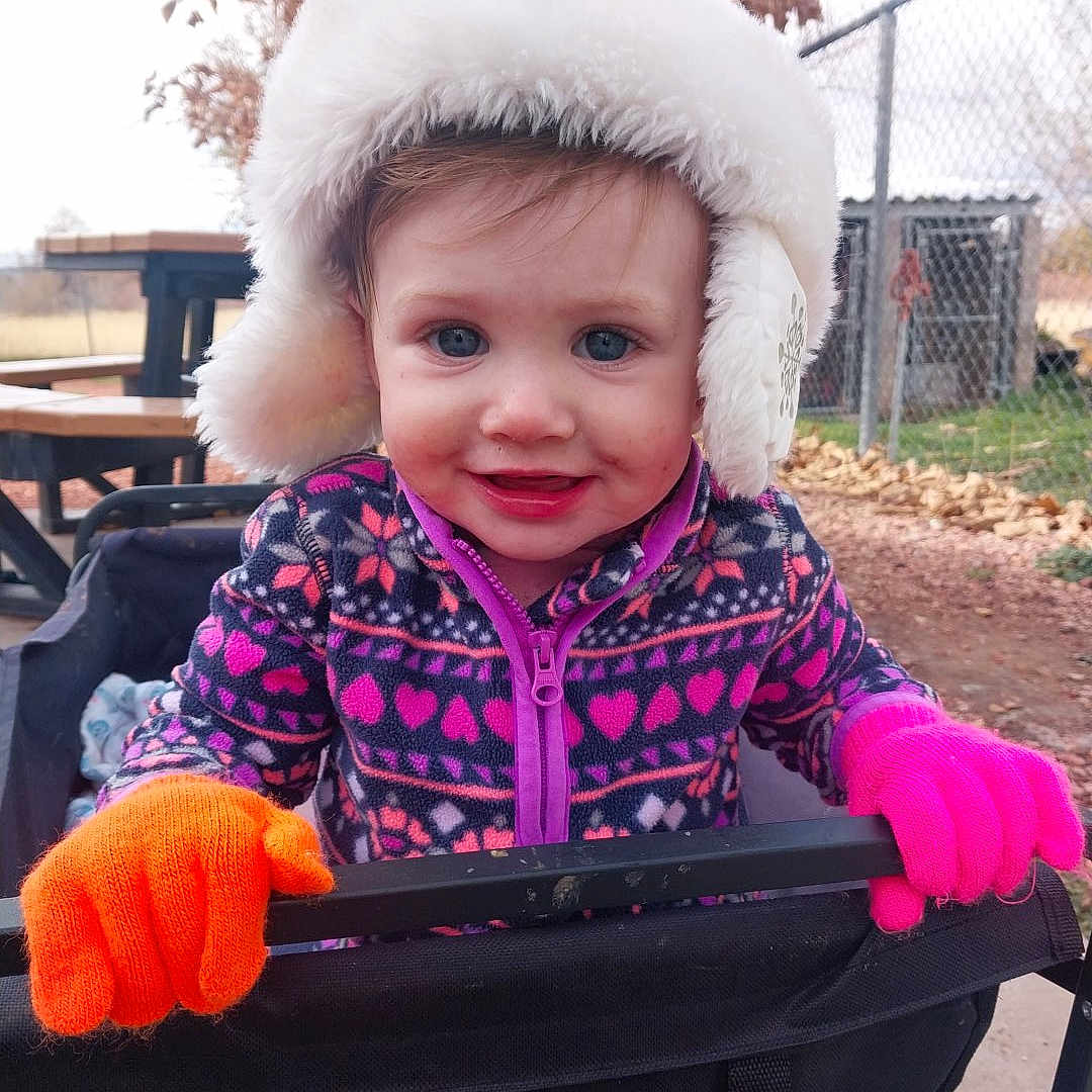 Journie is registered to the contest to win money with this photo: autumn, chain_link_fence, child, cold_weather, colorful_clothing, cute, face, gloves, jacket, leaves, outdoor, person, playful, portrait, smiling, stroller, toddler, wagon, winter_hat, young_child