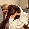 person, dog, puppy, chocolate_labrador, hug, couch, cat, glasses, bracelet, sweatshirt, indoor, cozy, affection, pet, animal, relaxation, home, furniture, window_blinds, blanket