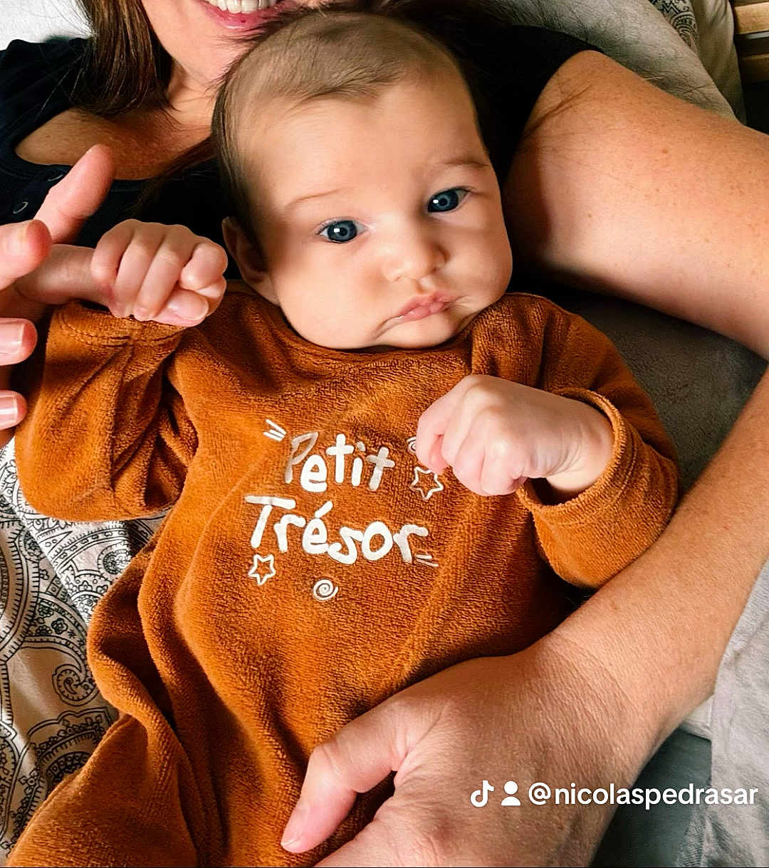 Noah a rejoint le concours — aidez-le/la à gagner de superbes lots ! baby, infant, child, person, holding, hand, finger, clothing, brown_outfit, text_on_clothing, face, blue_eyes, smile, adult, skin, closeup, portrait, cuddle, indoor, blanket