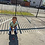Jace is registered to the contest to win money with this photo: child, boy, outdoor, sunlight, shadow, fence, pavement, casual_clothing, jeans, crocs, curly_hair, snack, daytime, residential_area, curious_expression, street, iron_fence, summer_shirt, casual_shoes, urban