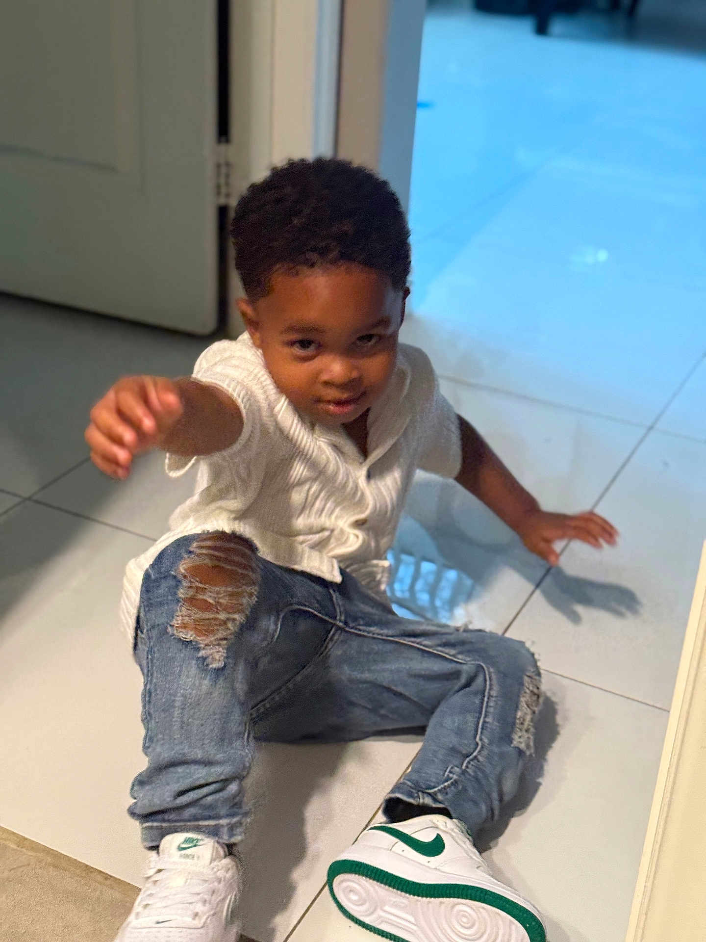 Jace is registered to the contest to win money with this photo: child, boy, indoor, floor, tile_floor, white_sweater, ripped_jeans, sneakers, nike, green, white, smiling, reaching, playing, casual_clothing, young_child, curly_hair, door, reflection, pose