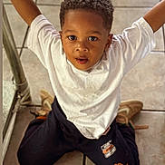 Jace joined the competition — help win amazing prizes! child, boy, kneeling, white_shirt, black_pants, orange_design, curly_hair, indoors, tile_floor, casual_clothing, shoes, arms_raised, looking_up, playful, young, portrait, expression, floor, person, cute