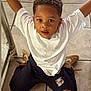 child, boy, kneeling, white_shirt, black_pants, orange_design, curly_hair, indoors, tile_floor, casual_clothing, shoes, arms_raised, looking_up, playful, young, portrait, expression, floor, person, cute