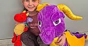 Léonie participe au concours pour gagner de l'argent avec cette photo : child, smiling, plush_toy, dragon, purple, red, yellow, indoor, tile_floor, crouching, happy, person, toy, casual_clothing, socks, wall, playful, cute, young, portrait
