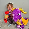 child, smiling, plush_toy, dragon, purple, red, yellow, indoor, tile_floor, crouching, happy, person, toy, casual_clothing, socks, wall, playful, cute, young, portrait