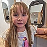 child, girl, blonde_hair, long_hair, tshirt, sequins, bird_design, indoor, mirror, reflection, wooden_counter, wall, casual_clothing, portrait, smiling, young_person, hair_bangs, room, home_interior, standing