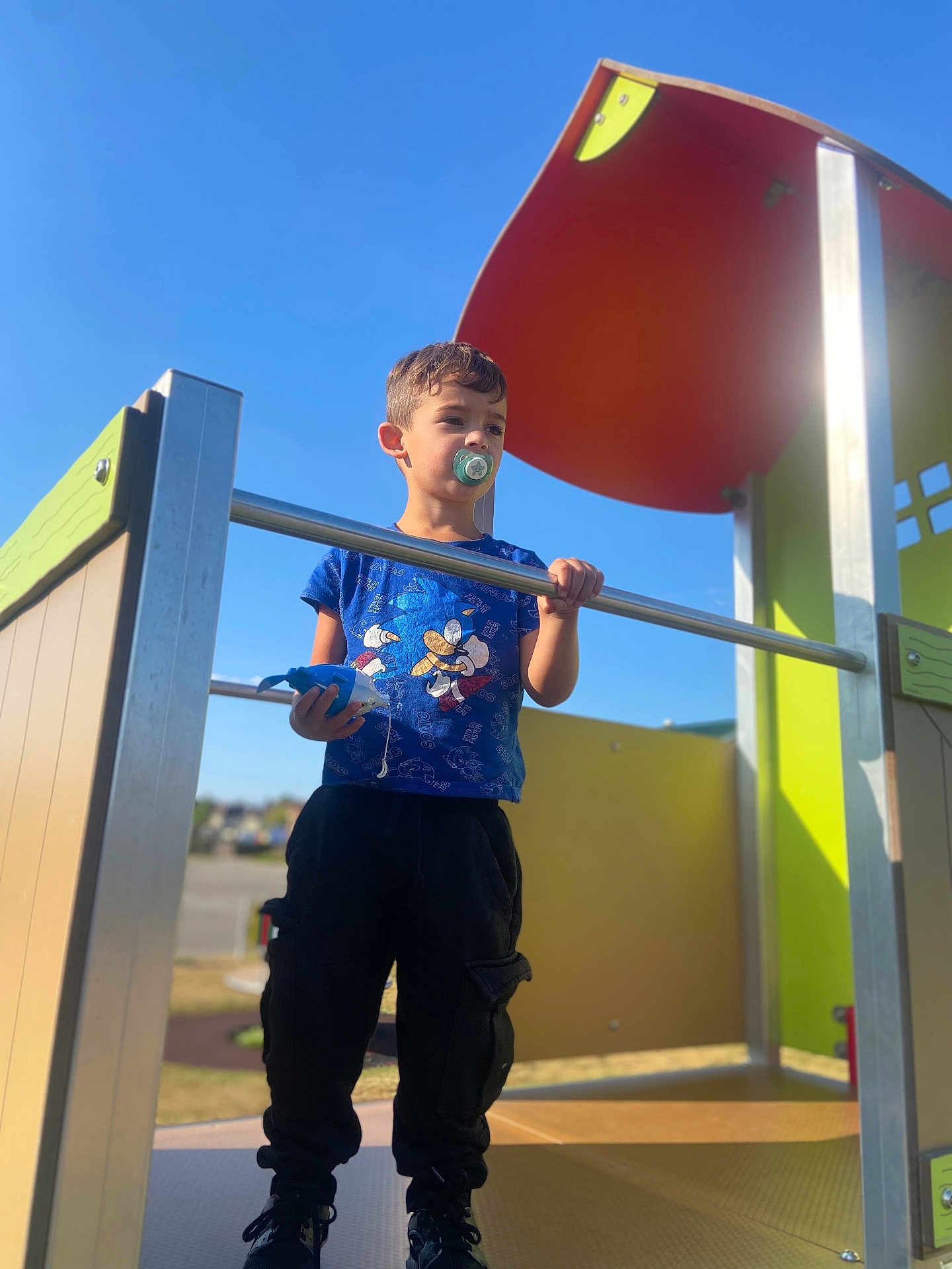 Rehan participe au concours pour gagner de l'argent avec cette photo : child, boy, playground, outdoor, toy, pacifier, blue_sky, tshirt, sonic_the_hedgehog, black_pants, sneakers, metal_rail, shade, sunlight, daytime, play_equipment, casual_clothing, young, standing, holding