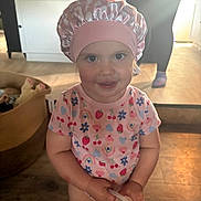 Amiyah is registered to the contest to win money with this photo: basket, cabinet, child, colorful_shirt, cute, face, flower_pattern, fruit_pattern, holding_object, home, indoor, lighting, person_in_background, pink_bonnet, playful, portrait, smiling, toddler, wooden_floor, young_child