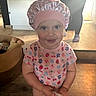 basket, cabinet, child, colorful_shirt, cute, face, flower_pattern, fruit_pattern, holding_object, home, indoor, lighting, person_in_background, pink_bonnet, playful, portrait, smiling, toddler, wooden_floor, young_child