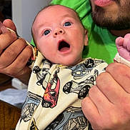 Benjamin is registered to the contest to win money with this photo: baby, man, adult, child, onesie, car_pattern, beard, green_shirt, hands, indoor, table, wood, surprise, expression, person, face, holding, curious, clothing, portrait